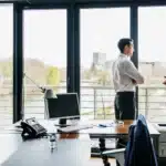 A photo of an office space with 2 men talking next to the window.